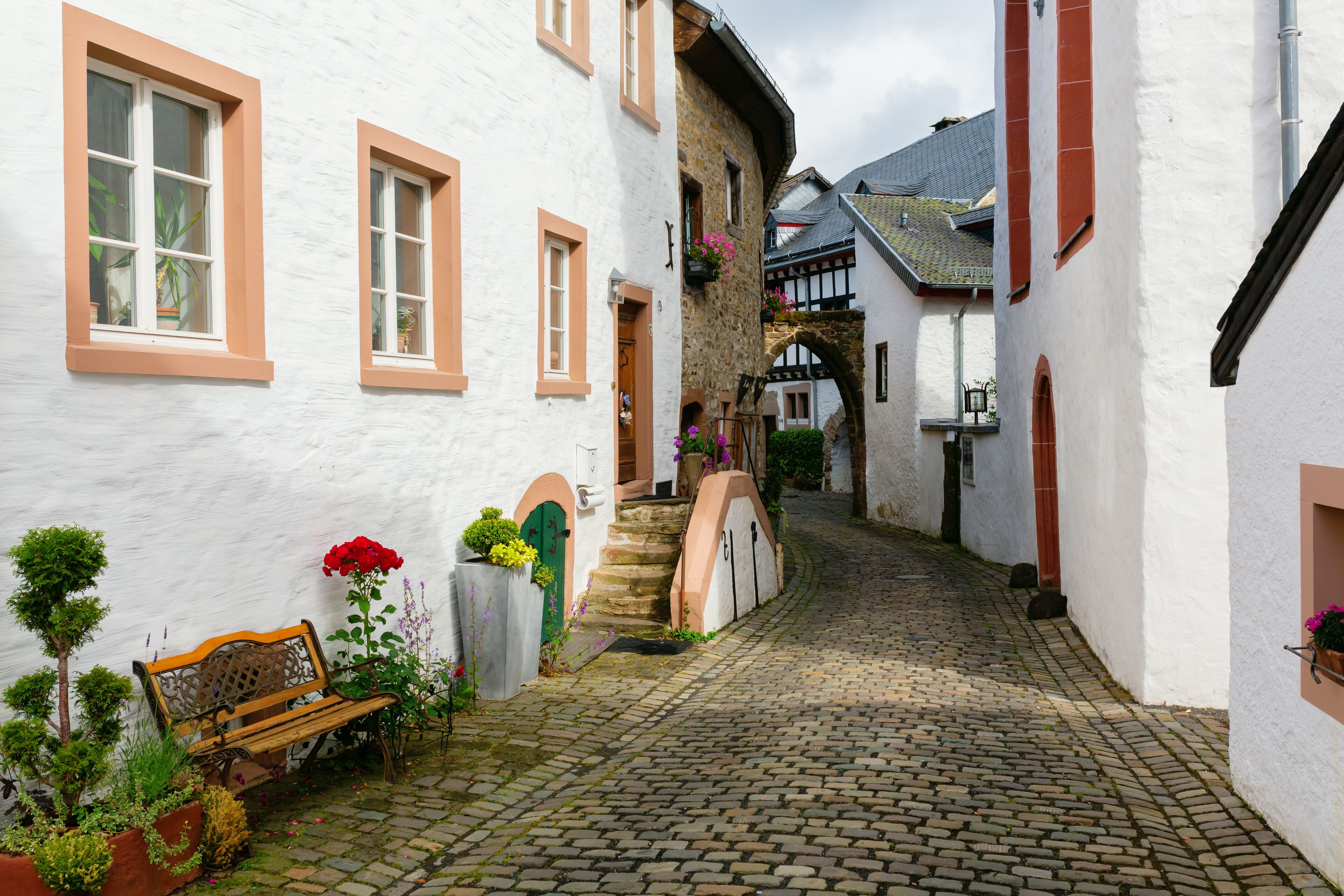 A cobblestone street in a European village with white buildings, a wooden bench, and potted plants.