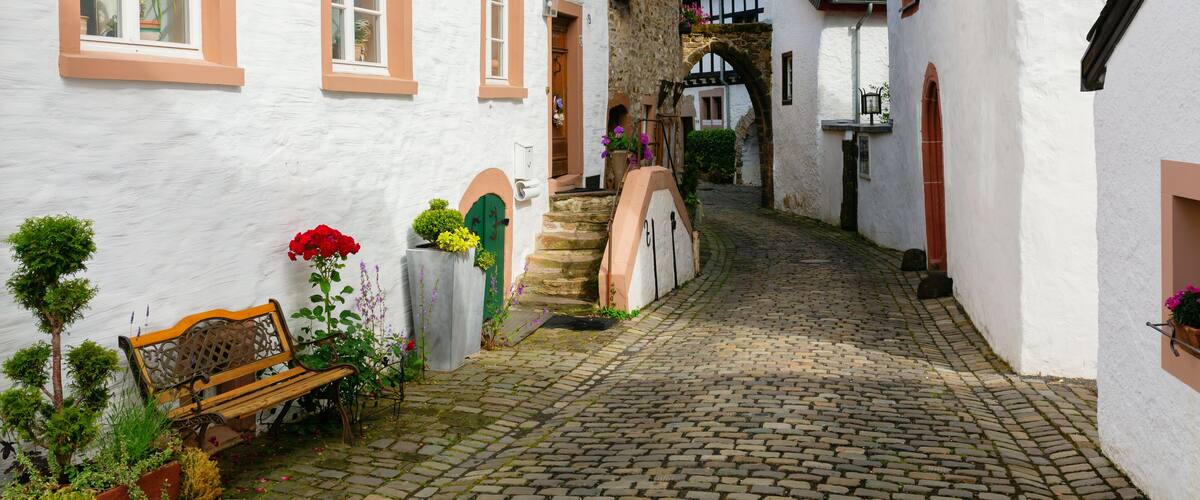 A cobblestone street in a European village with white buildings, a wooden bench, and potted plants.