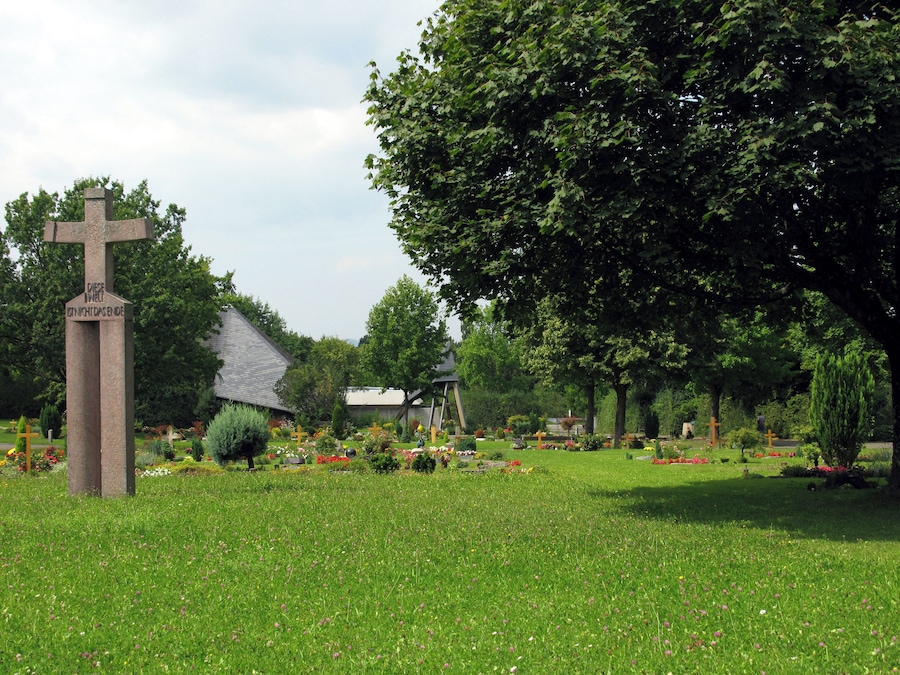 Een begraafplaats met een groot kruismonument, omringd door bomen en kleurrijke bloemen.
