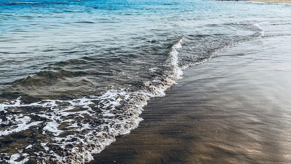 A beach with gentle waves, clear blue sky, and distant rocky outcrops.