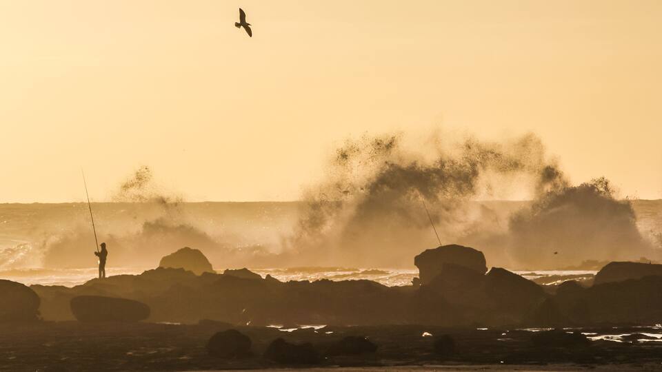 A person fishing by the sea with waves crashing against rocks.