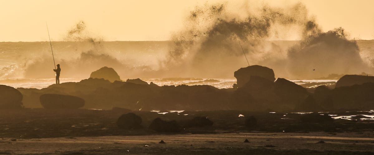 A person fishing by the sea with waves crashing against rocks.