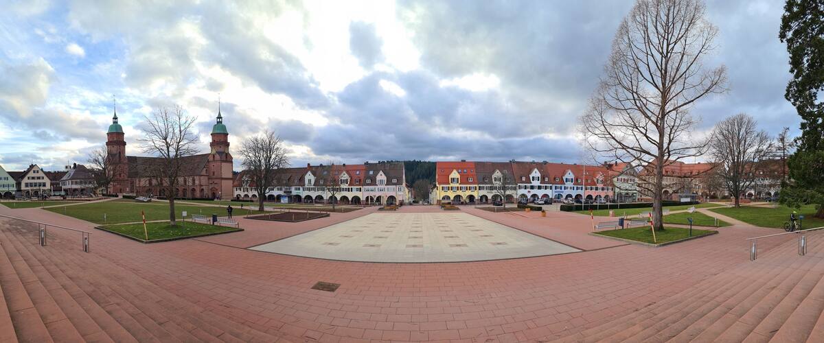 A large open square with a central plaza, surrounded by historic buildings with red roofs and a prominent tower.