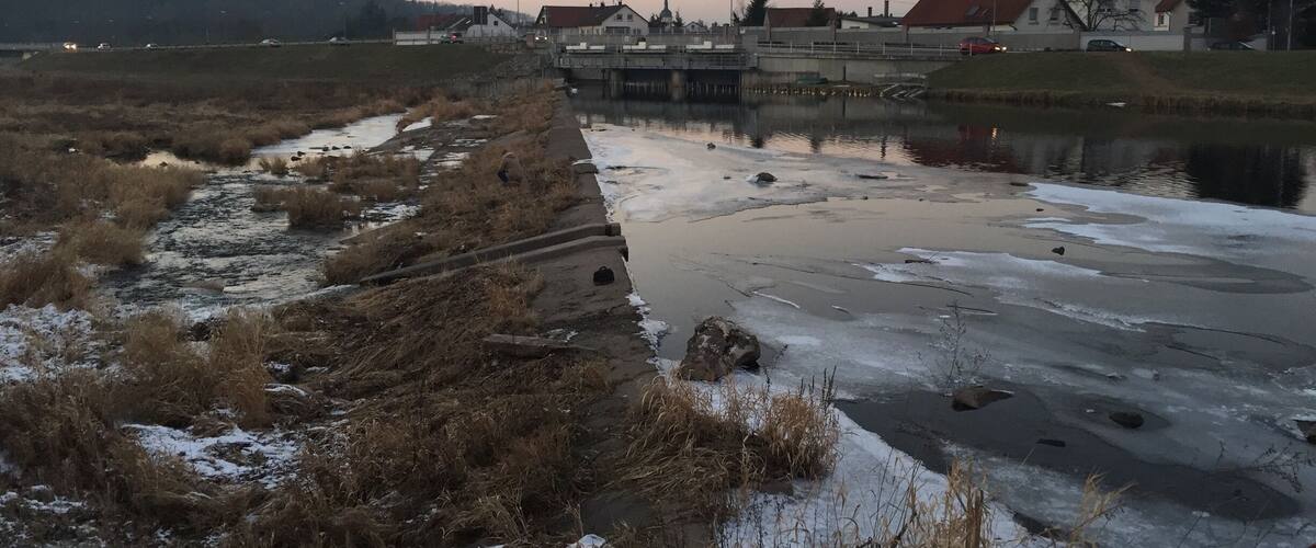 A frozen river with a concrete embankment, surrounded by grassy areas and residential buildings.