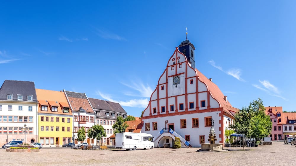Une place historique, bordée de bâtiments aux couleurs vives, avec une église imposante et un fourgon blanc garé au centre.