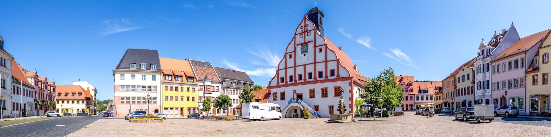 A historic town square with colorful buildings, a prominent church, and a white van parked in the center.