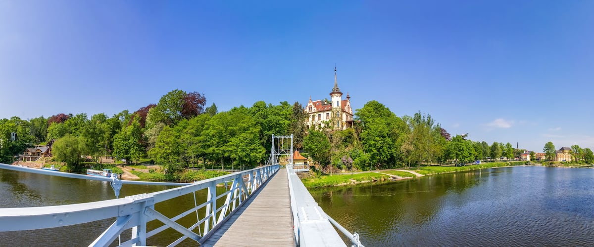 A wooden bridge over a river, surrounded by lush greenery and historic buildings.