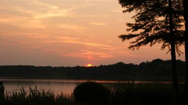 A sunset over a calm lake with silhouettes of trees and a person in the foreground.