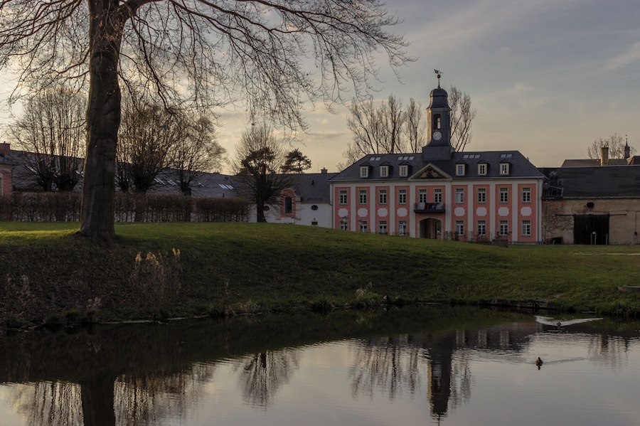 Un bâtiment historique doté d’un clocher, un bassin d’eau qui reflète sa façade, et un espace verdoyant avec des arbres.