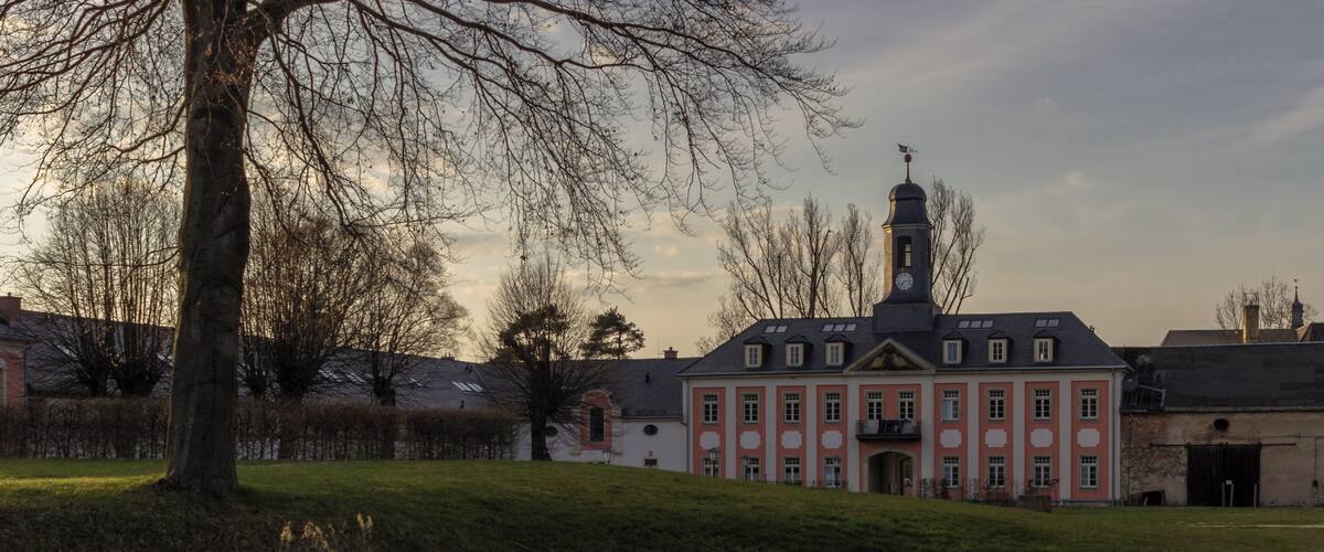 A historic building with a clock tower, a pond reflecting the structure, and a grassy area with trees.