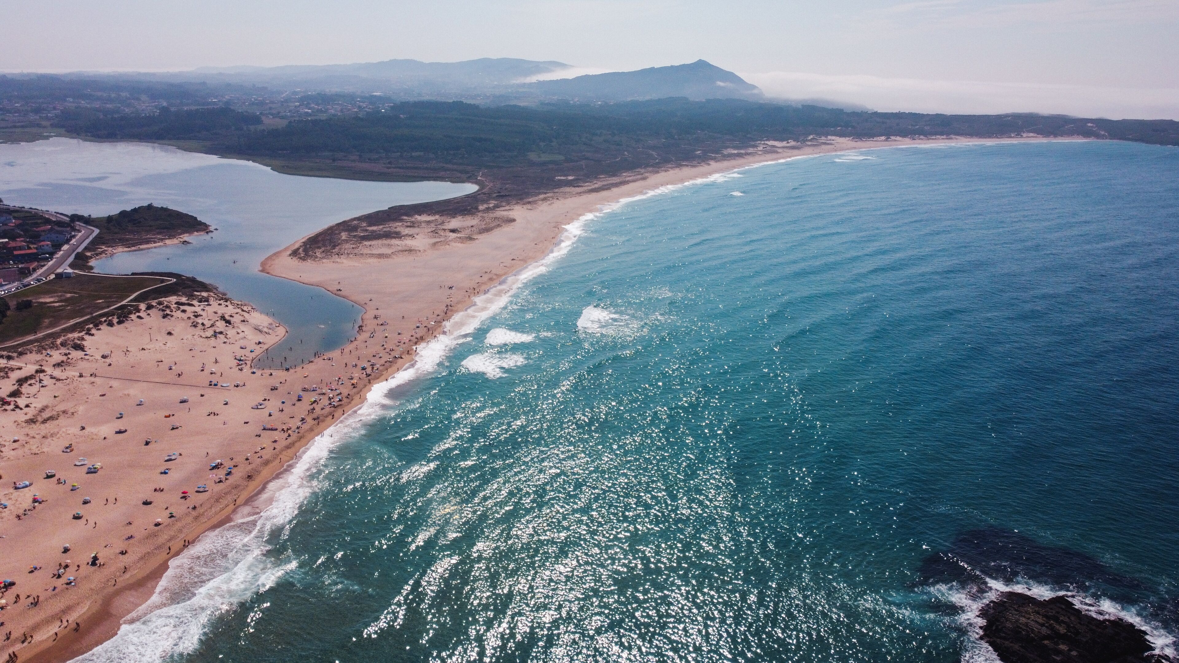 A coastal landscape with a sandy beach, a calm sea, and a mountainous backdrop.