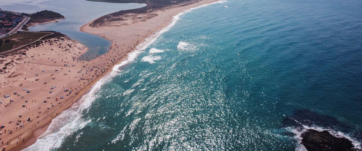 A coastal landscape with a sandy beach, a calm sea, and a mountainous backdrop.