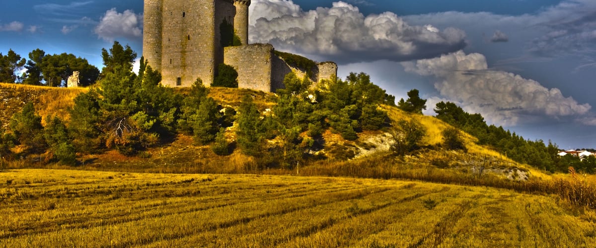 A medieval castle on a hill with surrounding fields and a partly cloudy sky.