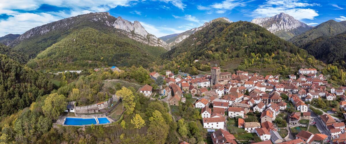 Un pueblo en la montaña con piscina, rodeado de colinas boscosas y una iglesia destacada.