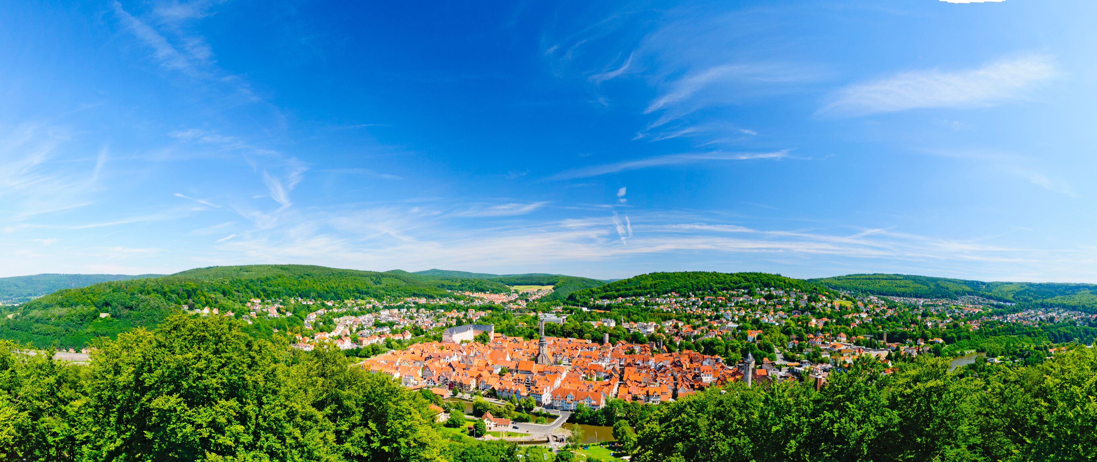 A panoramic view of a town nestled in a valley with a river, surrounded by lush green hills and a clear blue sky.