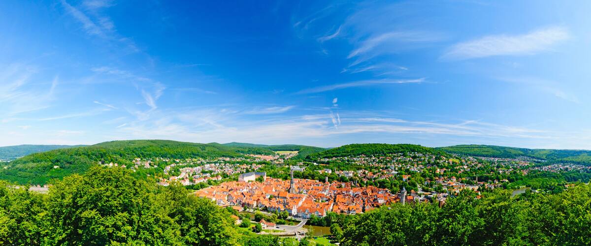 A panoramic view of a town nestled in a valley with a river, surrounded by lush green hills and a clear blue sky.