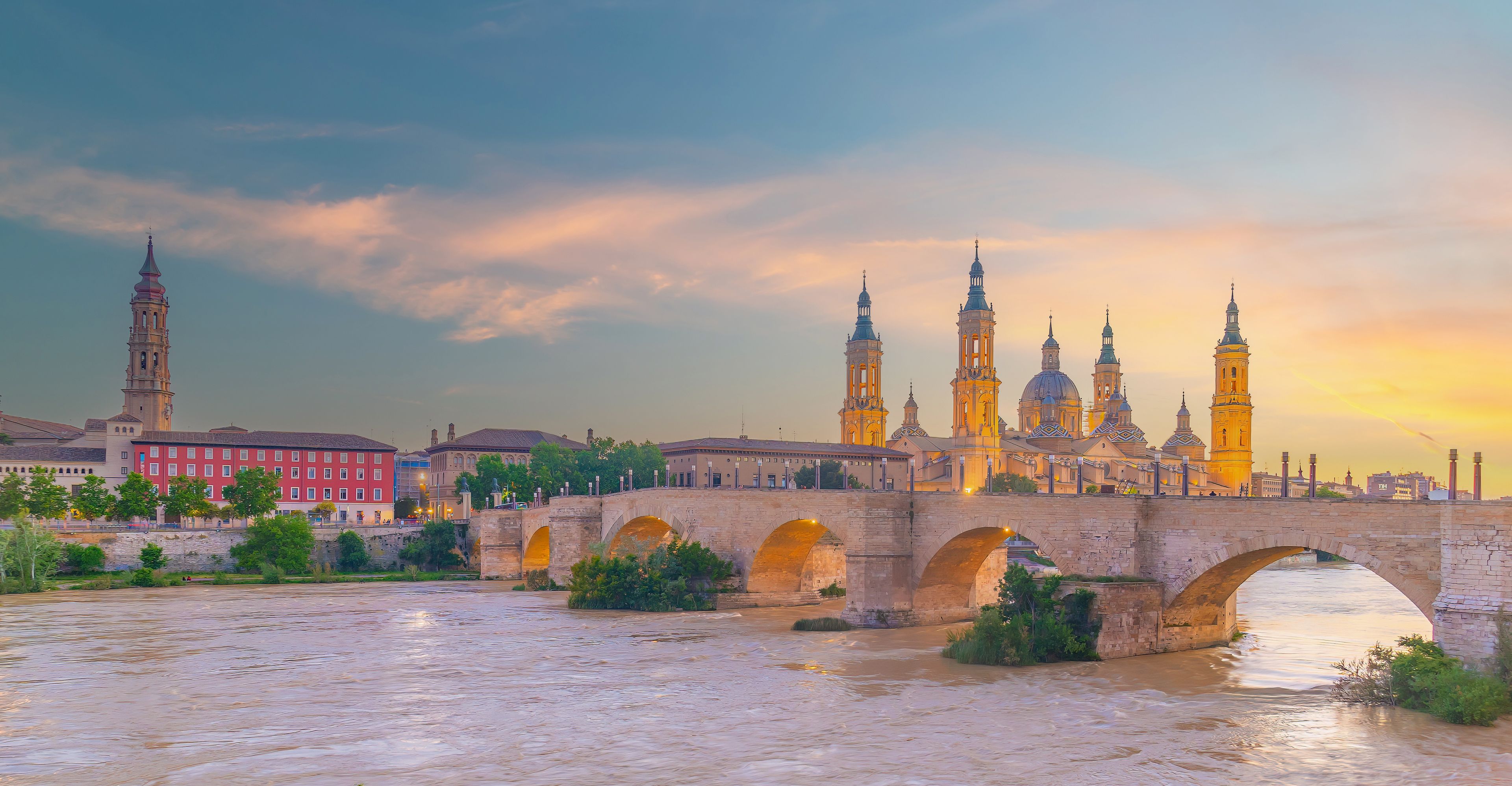 Une rivière traversée par un pont de pierre, un bâtiment rouge et une cathédrale dotée de plusieurs tours.