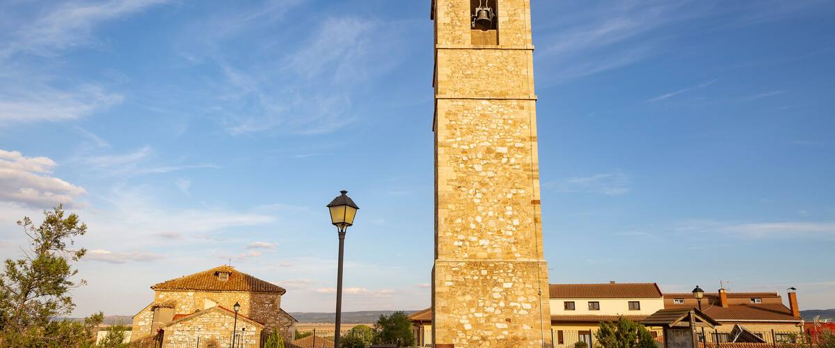 A tall stone tower with a bell, surrounded by houses and greenery under a clear sky.