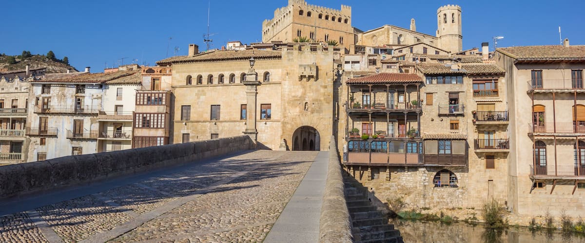 A historic town with a prominent castle, cobblestone streets, and a calm river reflecting the buildings.