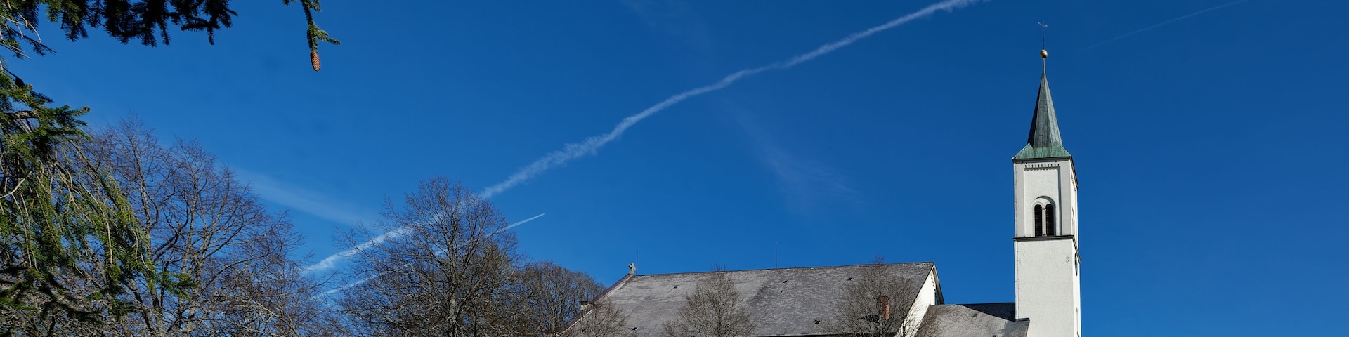 Una iglesia con un campanario alto y una cruz en la parte superior, rodeada de árboles y un cielo azul despejado.