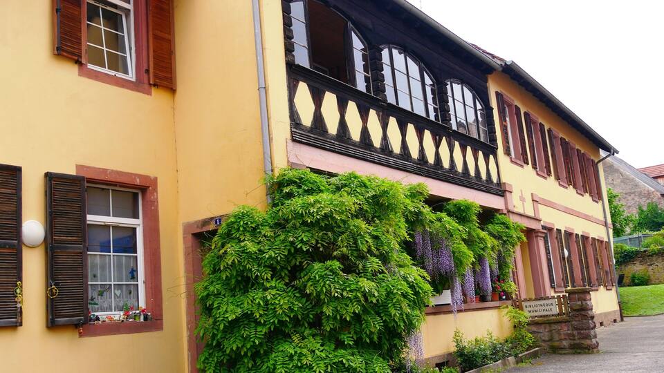 A yellow building with wooden shutters, a balcony with black balustrades, and green ivy climbing the walls.