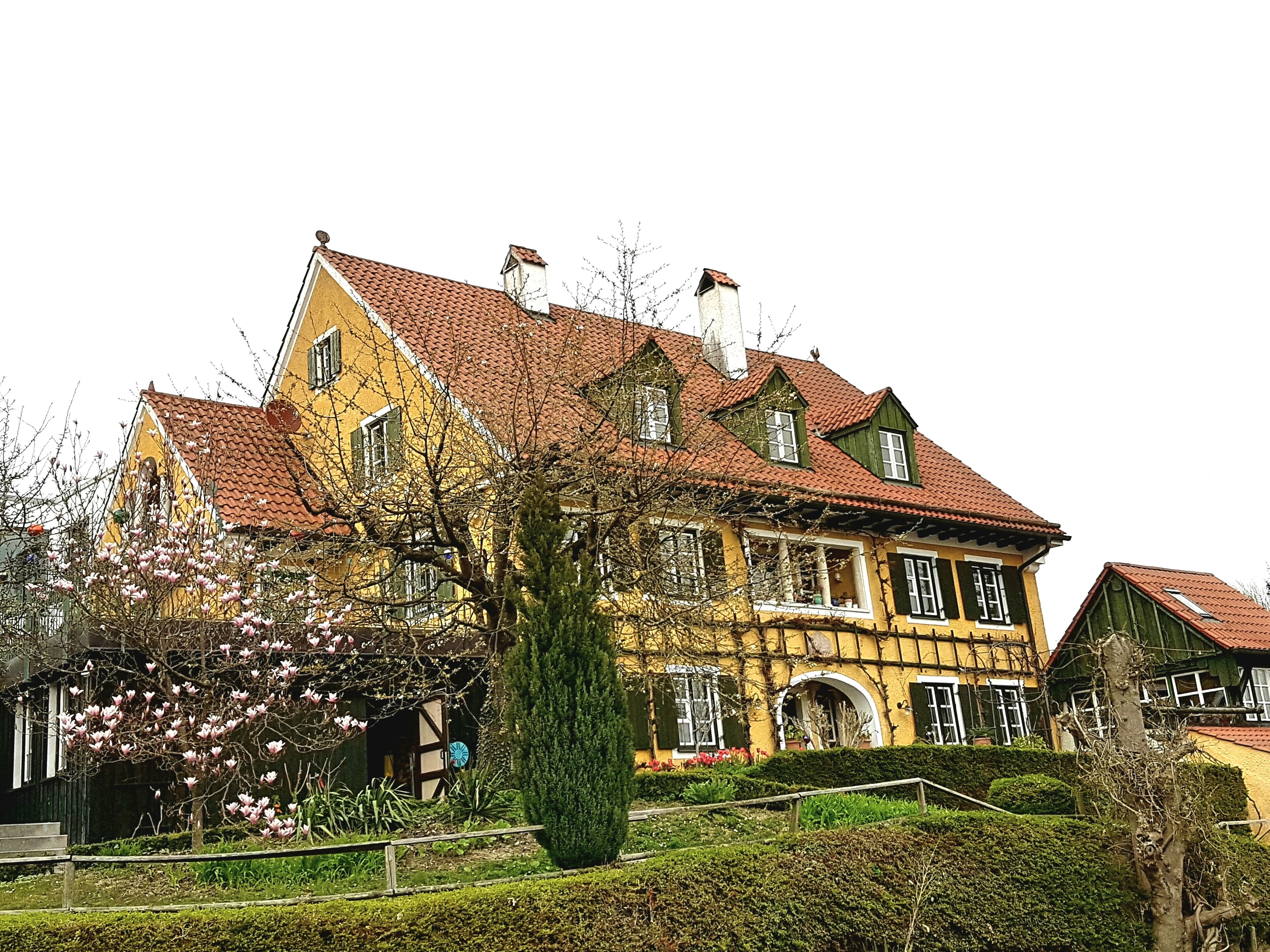 A large yellow building with a red-tiled roof, surrounded by greenery and flowering trees.