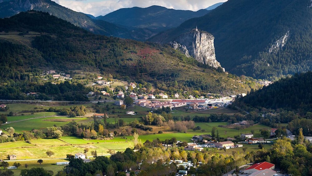 A mountainous landscape with a village nestled in a valley, a prominent castle-like structure on a cliff, and rolling green fields.