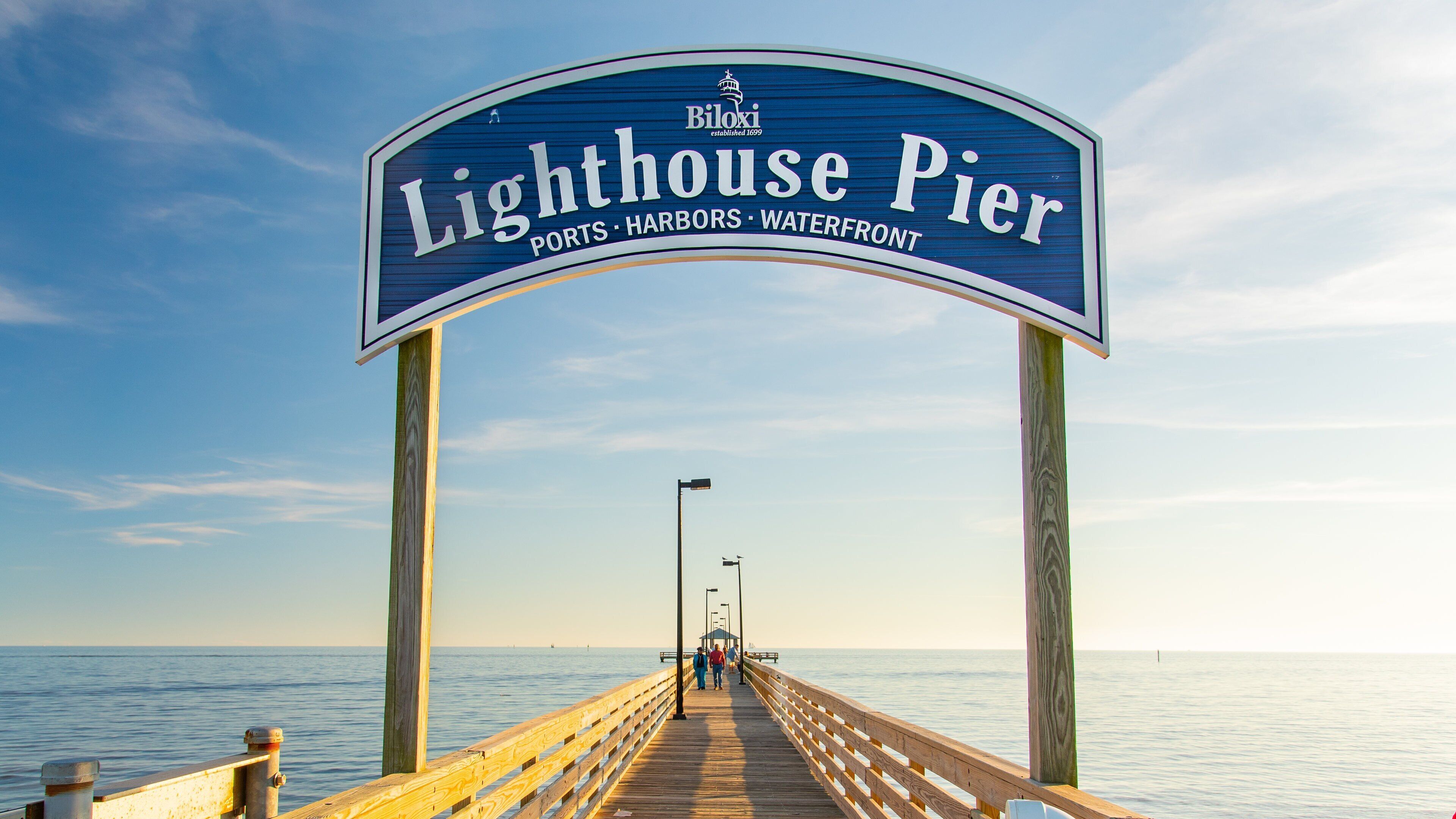 A wooden pier leading to a sign reading "Lighthouse Pier" with a clear sky and calm water in the background.