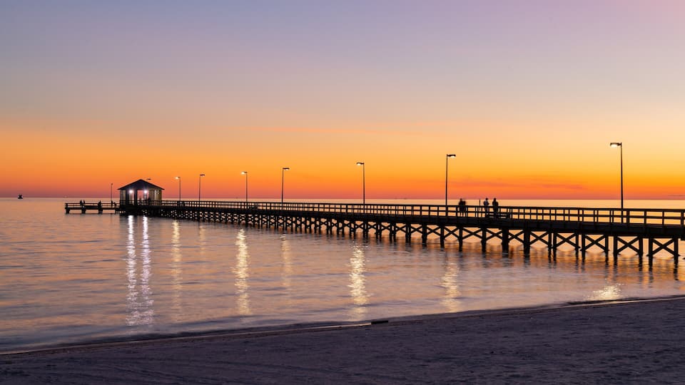 A pier extending into calm waters at sunset with a small shelter and street lamps.