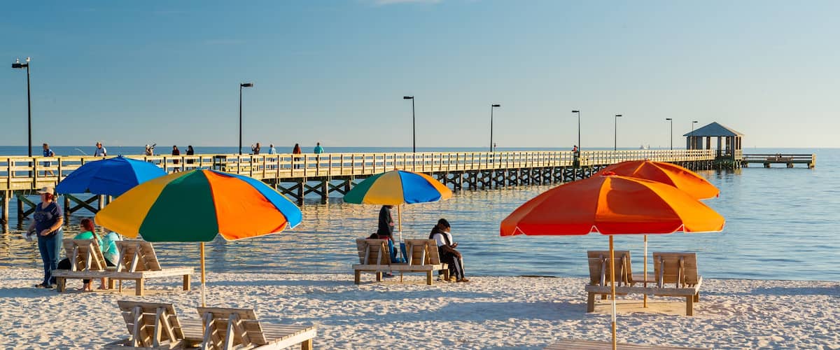 A beach with wooden chairs and colorful umbrellas, a pier extending into the water, and a lifeguard tower in the background.
