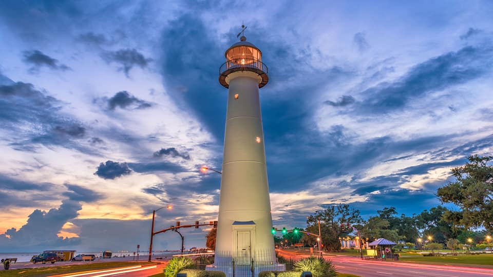 A tall white lighthouse with a balcony, surrounded by a well-maintained grassy area, a road with light trails, and a backdrop of a cloudy sky.
