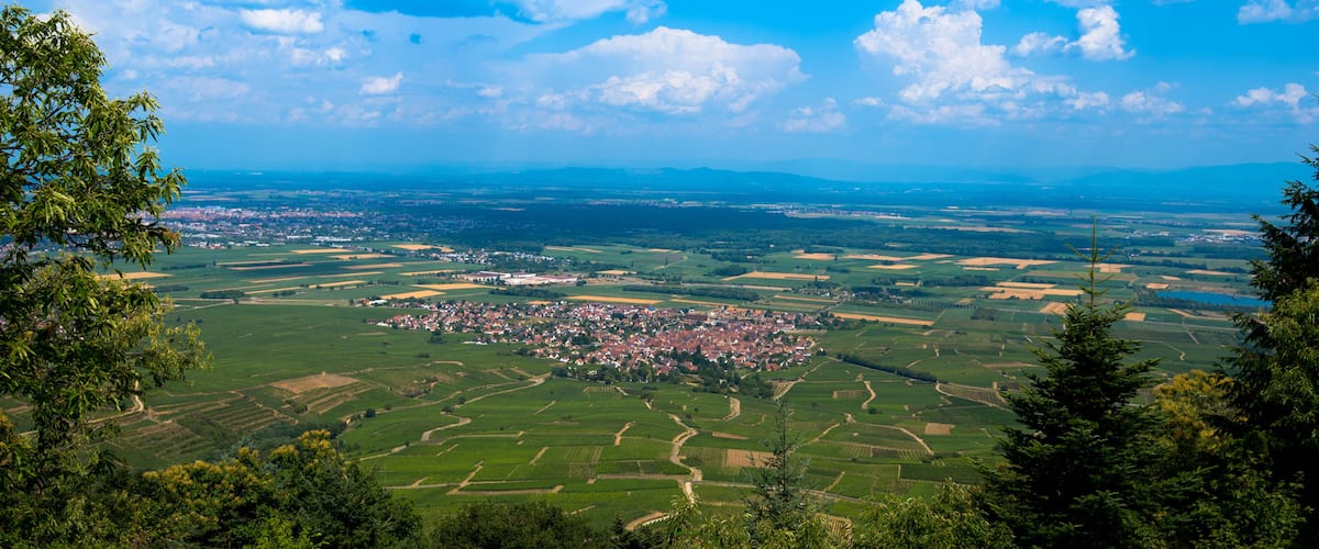 A panoramic view of a town surrounded by lush green fields and distant mountains under a clear blue sky.