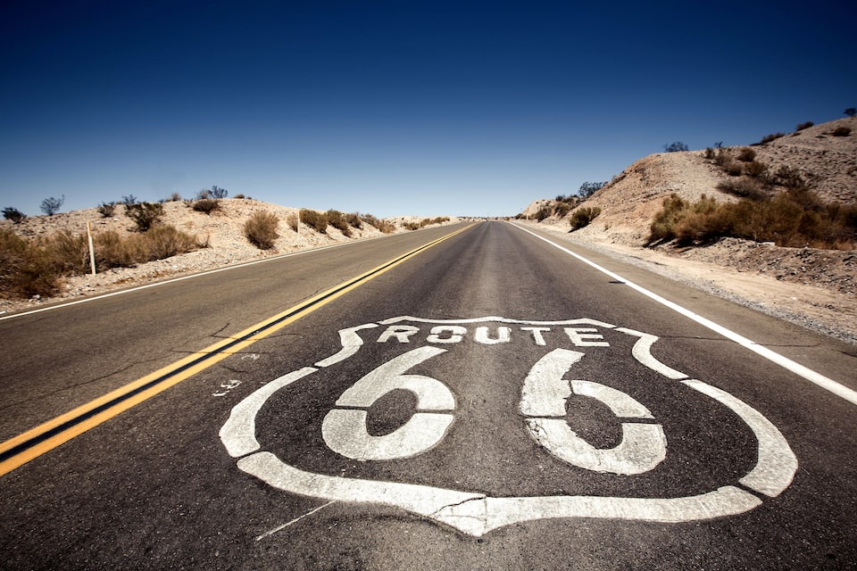 A road with Route 66 painted on the asphalt, surrounded by desert terrain and sparse vegetation.