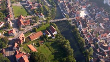 Vista aérea de un pueblo con un río que lo atraviesa, rodeado de campos verdes y casas con tejados rojos.