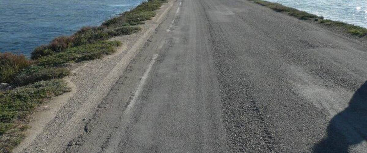 A gravel road alongside a body of water with distant land visible.