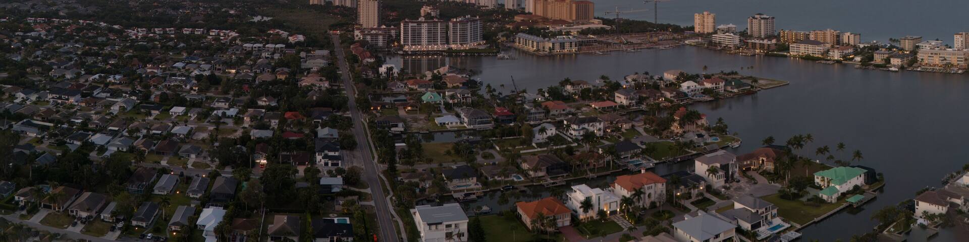 A coastal cityscape at sunset with a mix of residential and high-rise buildings, a marina, and a large body of water.