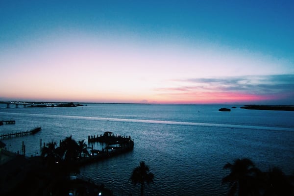 A sunset over a calm sea with a pier and palm trees.