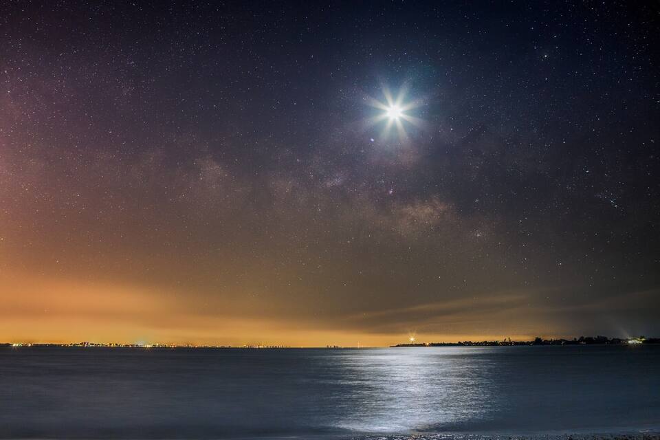 A starry night sky over a calm body of water with a bright star and a city skyline in the distance.