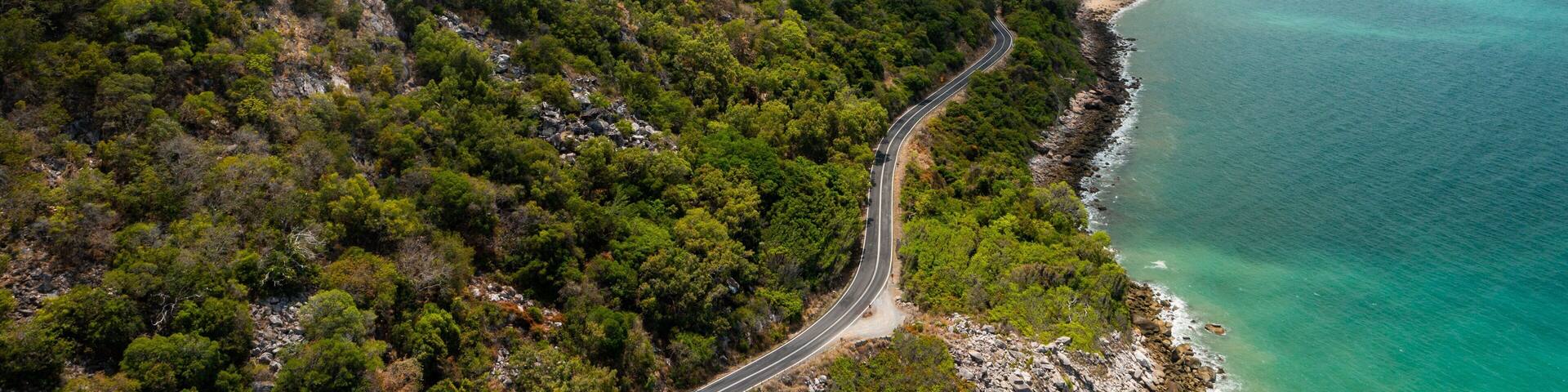 A winding road through a mountainous area with dense green forests, leading to a coastline with clear turquoise waters.