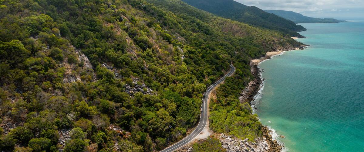A winding road through a mountainous area with dense green forests, leading to a coastline with clear turquoise waters.