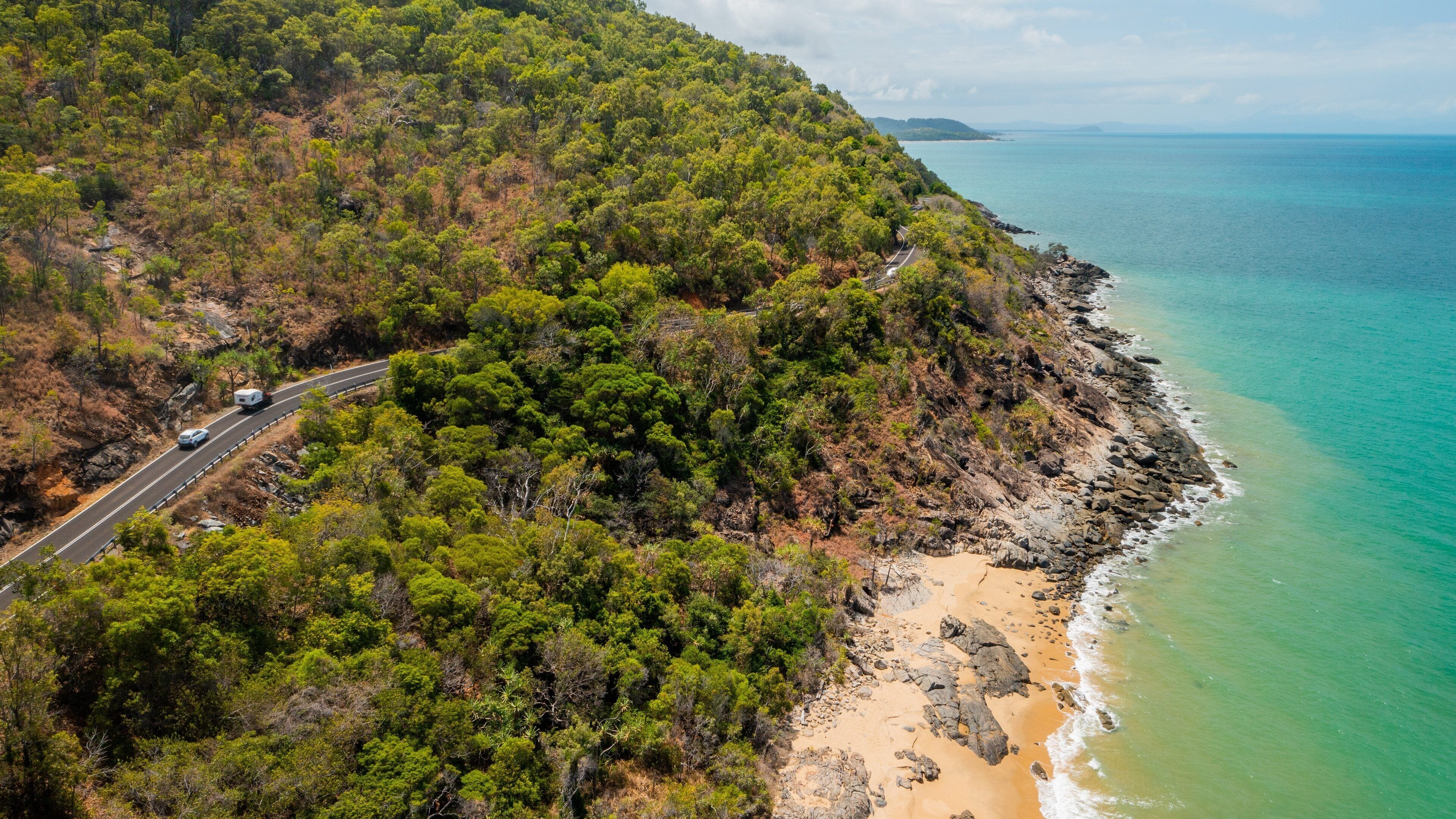 Uma estrada costeira serpenteia por uma floresta verde densa, levando a uma praia arenosa com águas turquesa cristalinas.