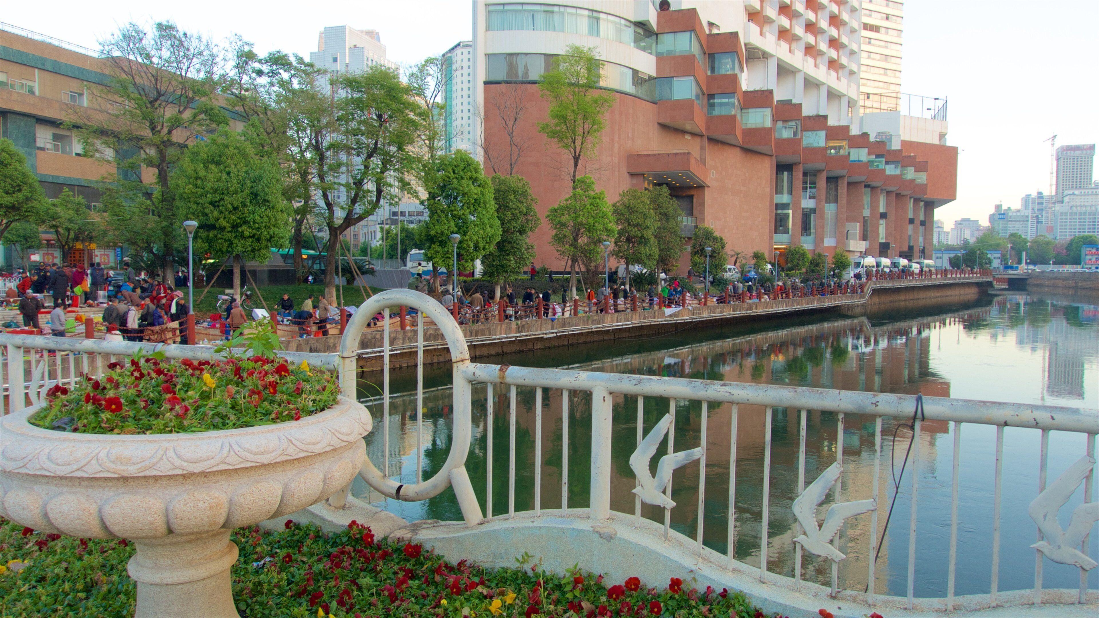 A riverside walkway with people, a bridge, and modern buildings in the background.
