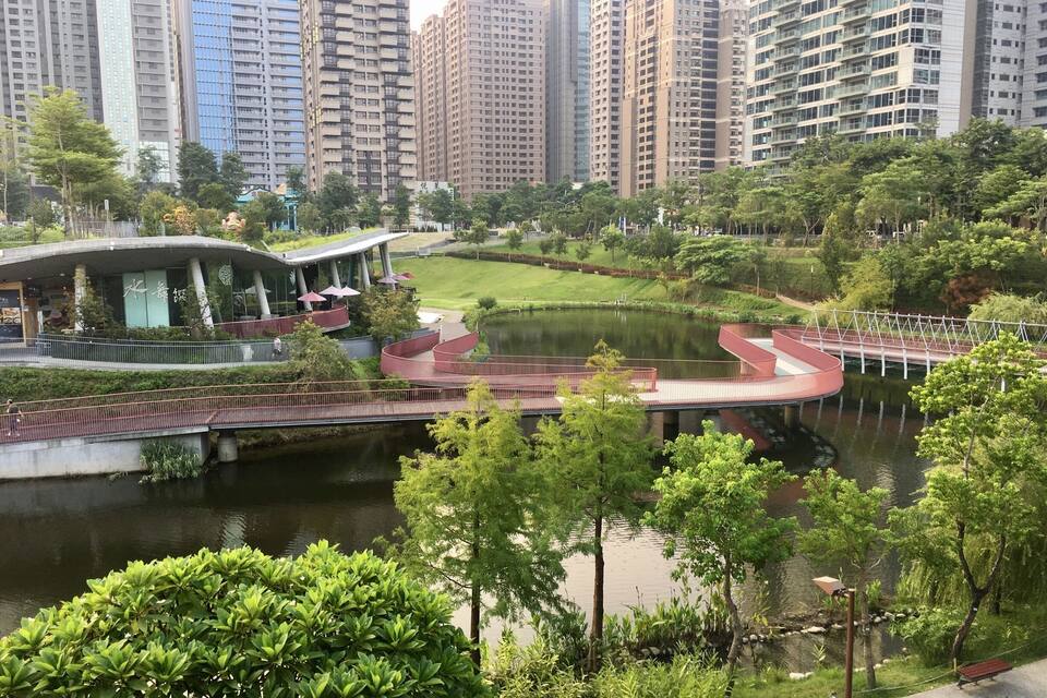 A riverside park with a curved bridge, lush greenery, and modern high-rise buildings in the background.