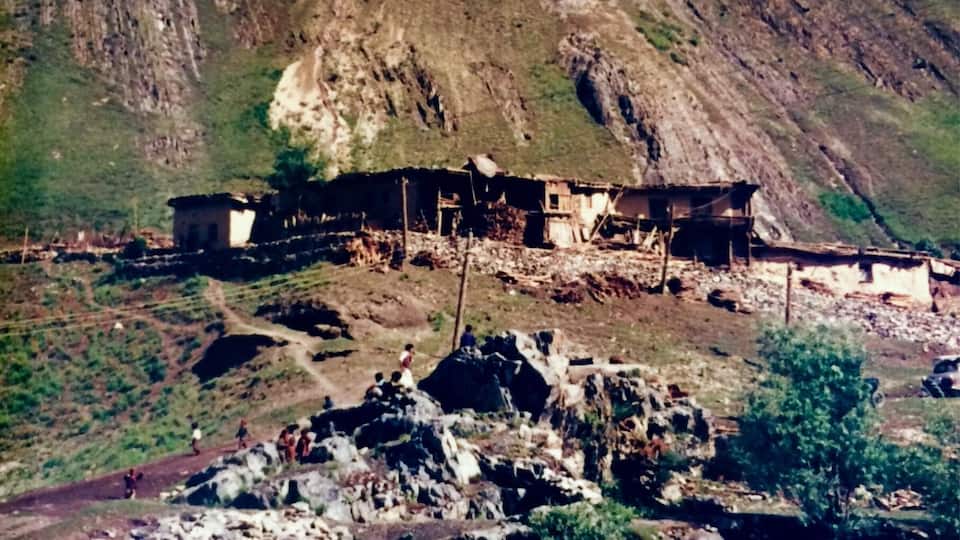 A mountainous landscape with a river in the foreground, a small settlement with buildings, and a prominent peak in the background.
