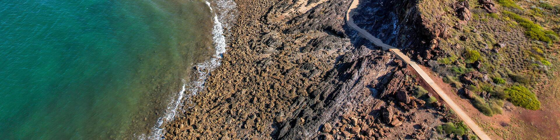 A coastal landscape with a rocky shoreline, a dirt path, and a distant view of a flat terrain.