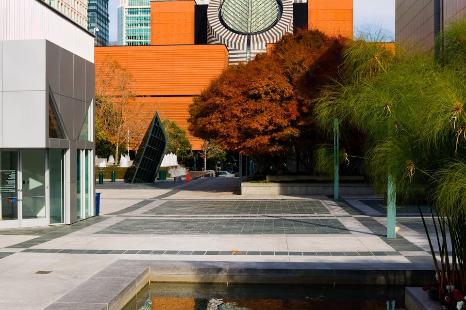 A modern urban plaza with a reflective pool, tall buildings, and a distinctive orange building with a circular design.