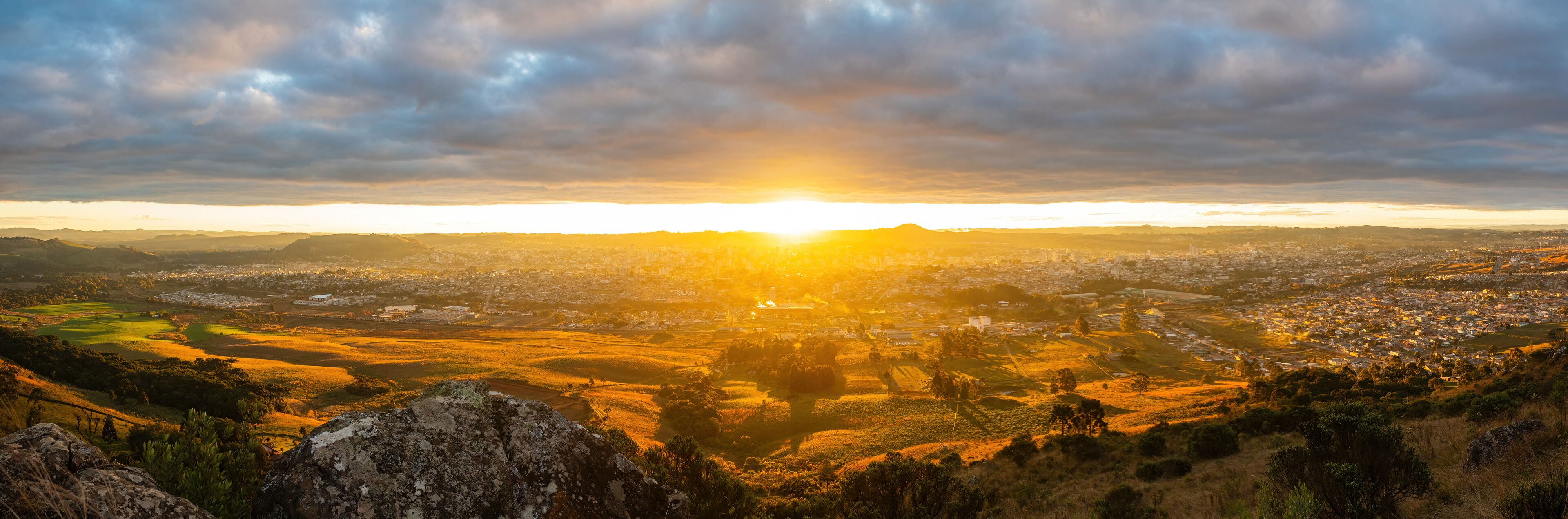 A panoramic view of a city at sunset with a prominent mountain in the foreground and a cloudy sky.