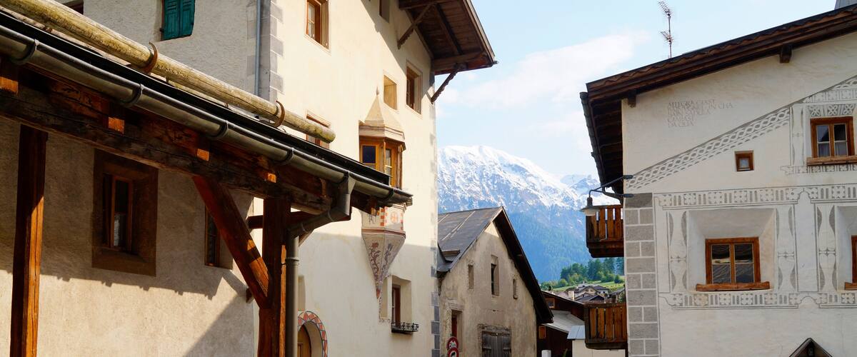 A narrow street in a mountainous village with traditional buildings and a snow-capped mountain in the background.