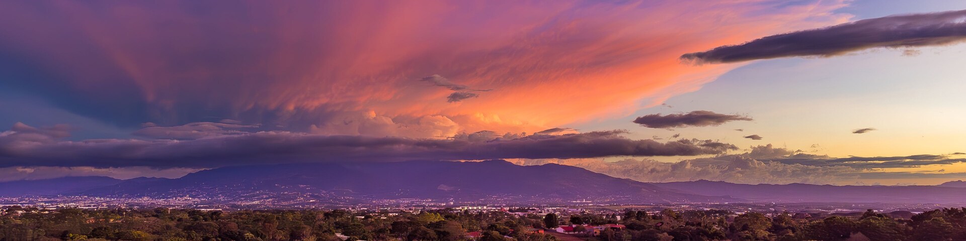 Un atardecer sobre una ciudad, con montañas al fondo y un bosque denso en primer plano.
