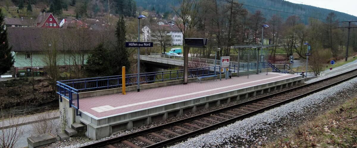 A train station platform with a sign reading "Höfen (Enz Nord)" and a bridge over tracks.